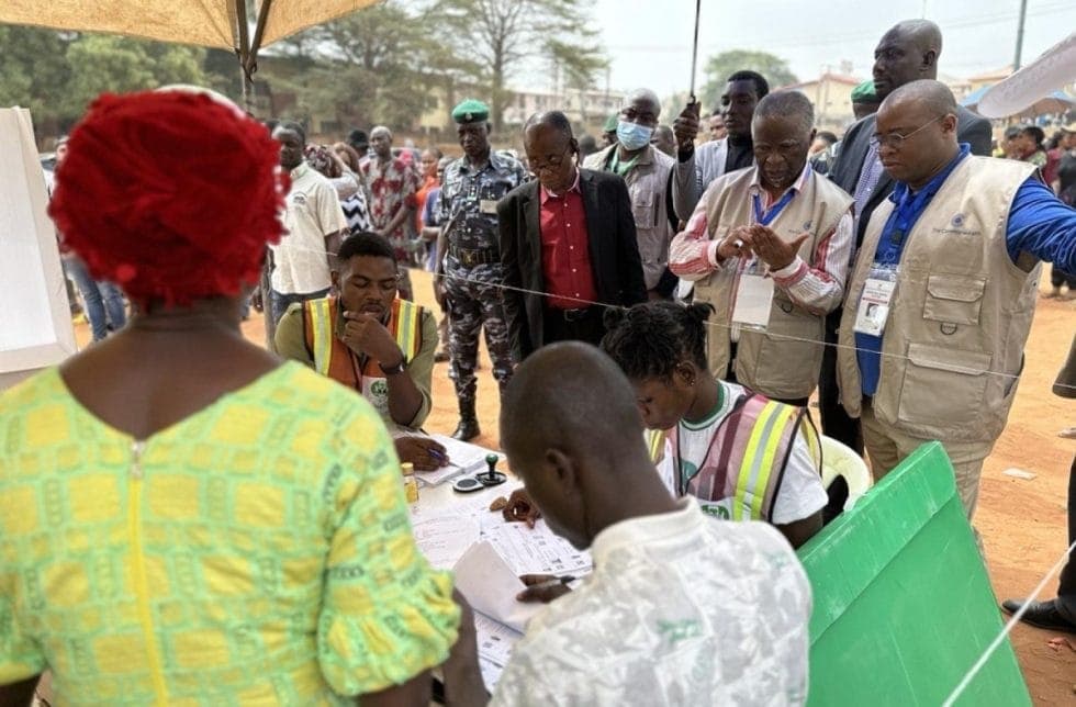 Voters and accredited observers gathered around an INEC polling table under a canopy, with a police officer and a green ballot box beside them.