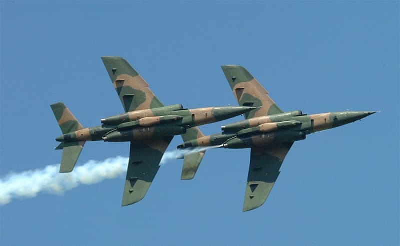 Two Nigerian Air Force fighter jets in camouflage livery flying in close formation against a clear sky, one trailing white smoke.