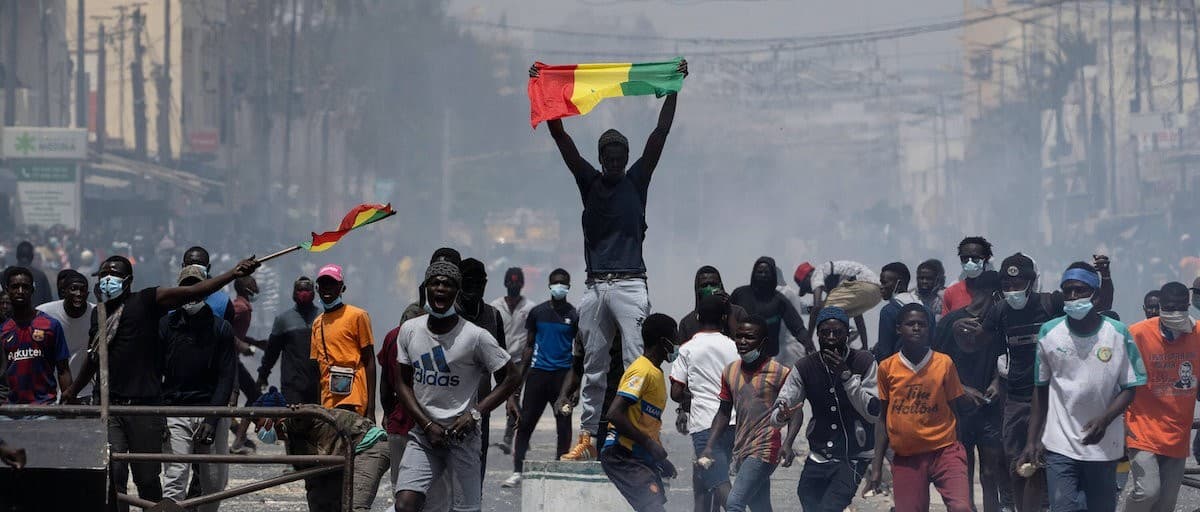 A demonstrator standing above a crowd in Dakar holding the Senegalese flag aloft, with protesters massed behind a barricade in the street.