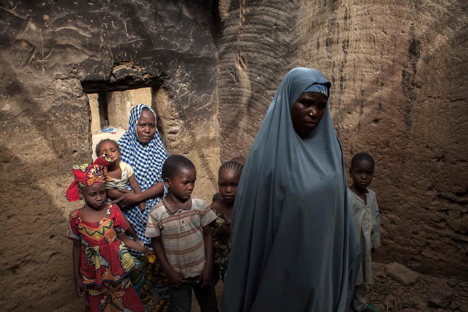 A woman in a pale blue hijab standing in a doorway of a stone building with six children gathered around her, one of them held in the arms of an older girl.