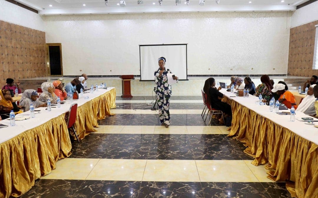 A speaker addressing a townhall from the centre of a U-shaped table arrangement, with participants seated along both sides of the room.