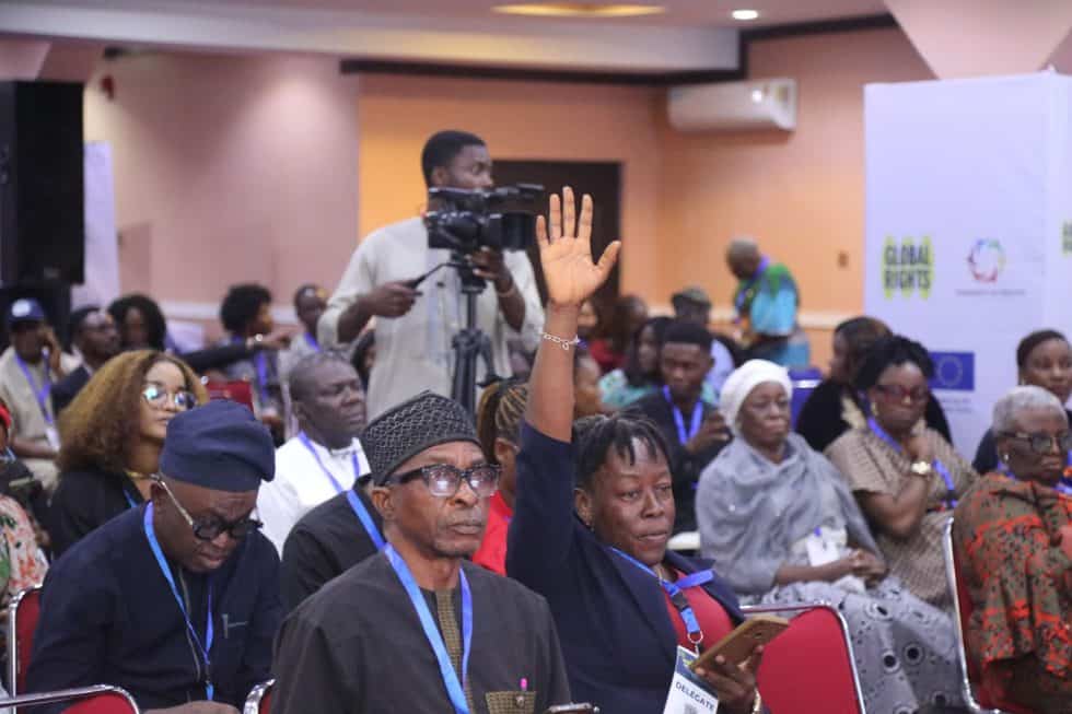 A delegate raising a hand to speak from the floor of a packed conference room, with a camera operator filming and a Global Rights banner behind the audience.