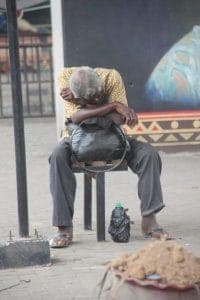 A man sitting on a low stool against a wall beside a bundle of belongings, on a Lagos street.