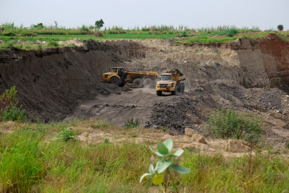 Two haul trucks working the floor of an open-cast mine pit, with green farmland along the rim above it.