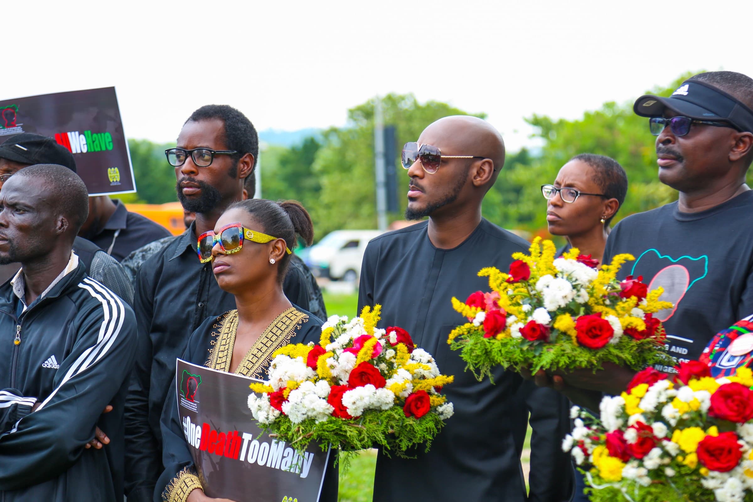 Mourners dressed in black at an outdoor memorial gathering, carrying wreaths of red, white and yellow flowers and placards reading “#OneDeathTooMany”.