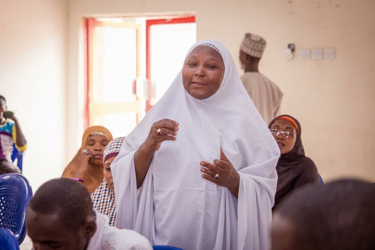 A woman standing to speak at a community meeting, addressing a seated room of women and men.