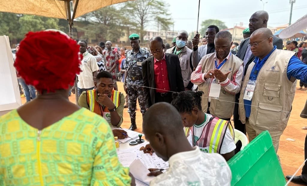 Voters and accredited observers gathered around an INEC polling table under a canopy, with a police officer and a green ballot box beside them.